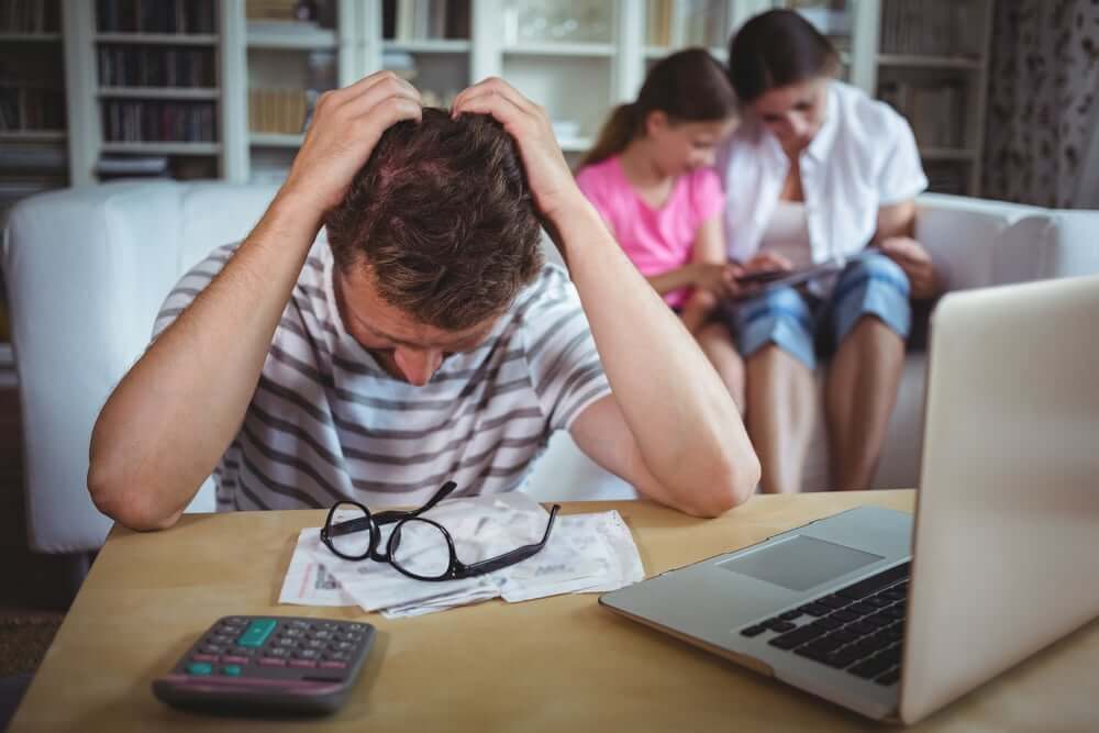 Worried man sitting at table with bills and laptop while his wife and daughter sitting on sofa-2